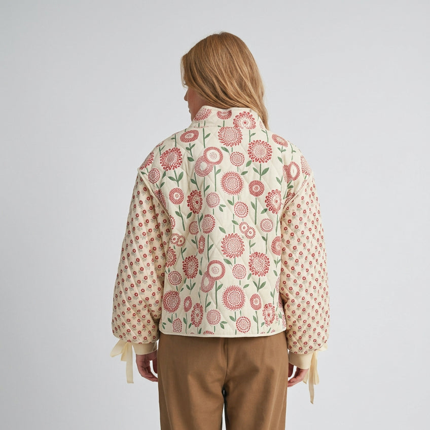 A woman shown from the back wearing a cropped jacket with a cream base, pink and red floral pattern on the back, and a pink polka-dot pattern on the sleeves and sides. She is also wearing brown pants against a plain beige background.