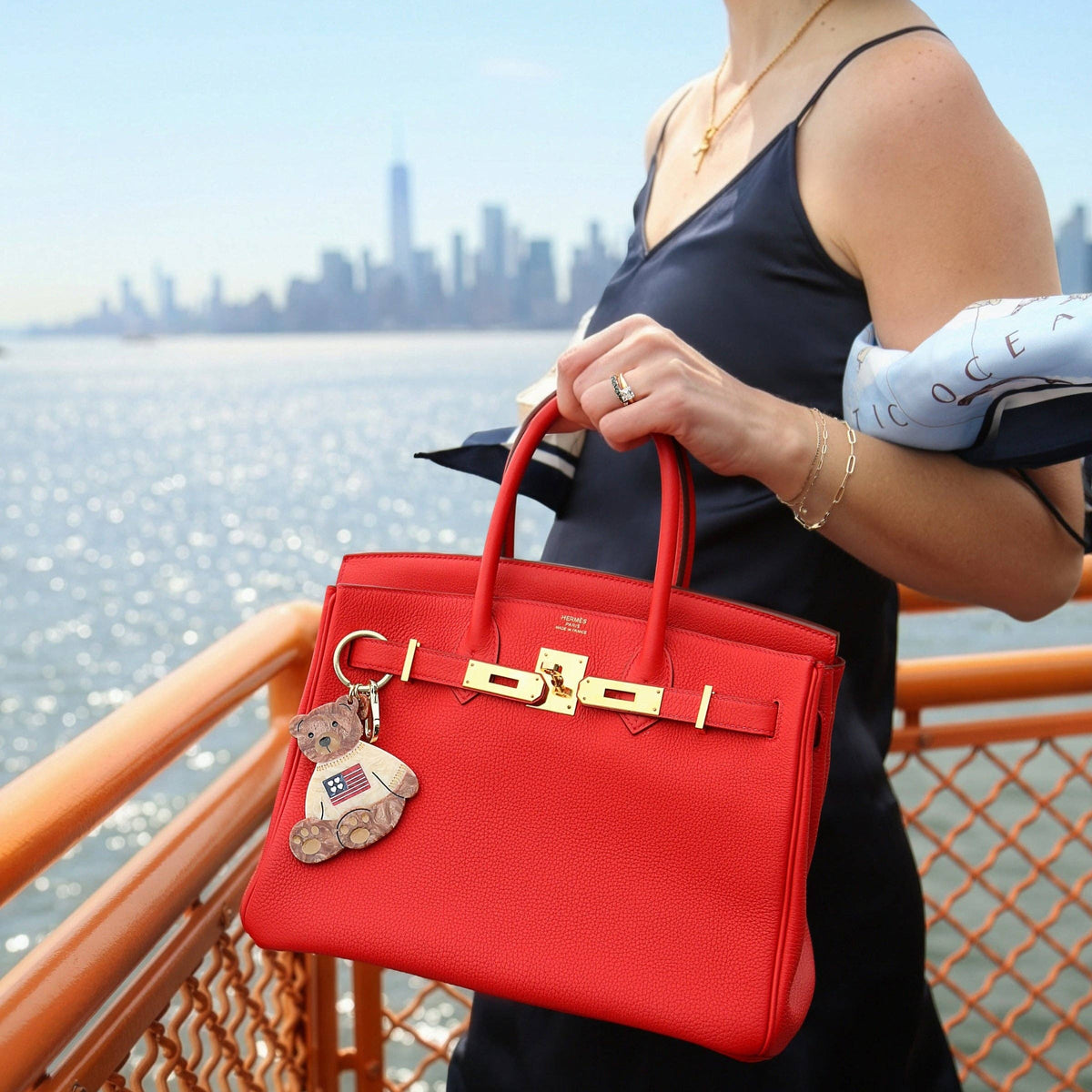 Person holding a red handbag with a teddy bear charm by a waterfront with city skyline.