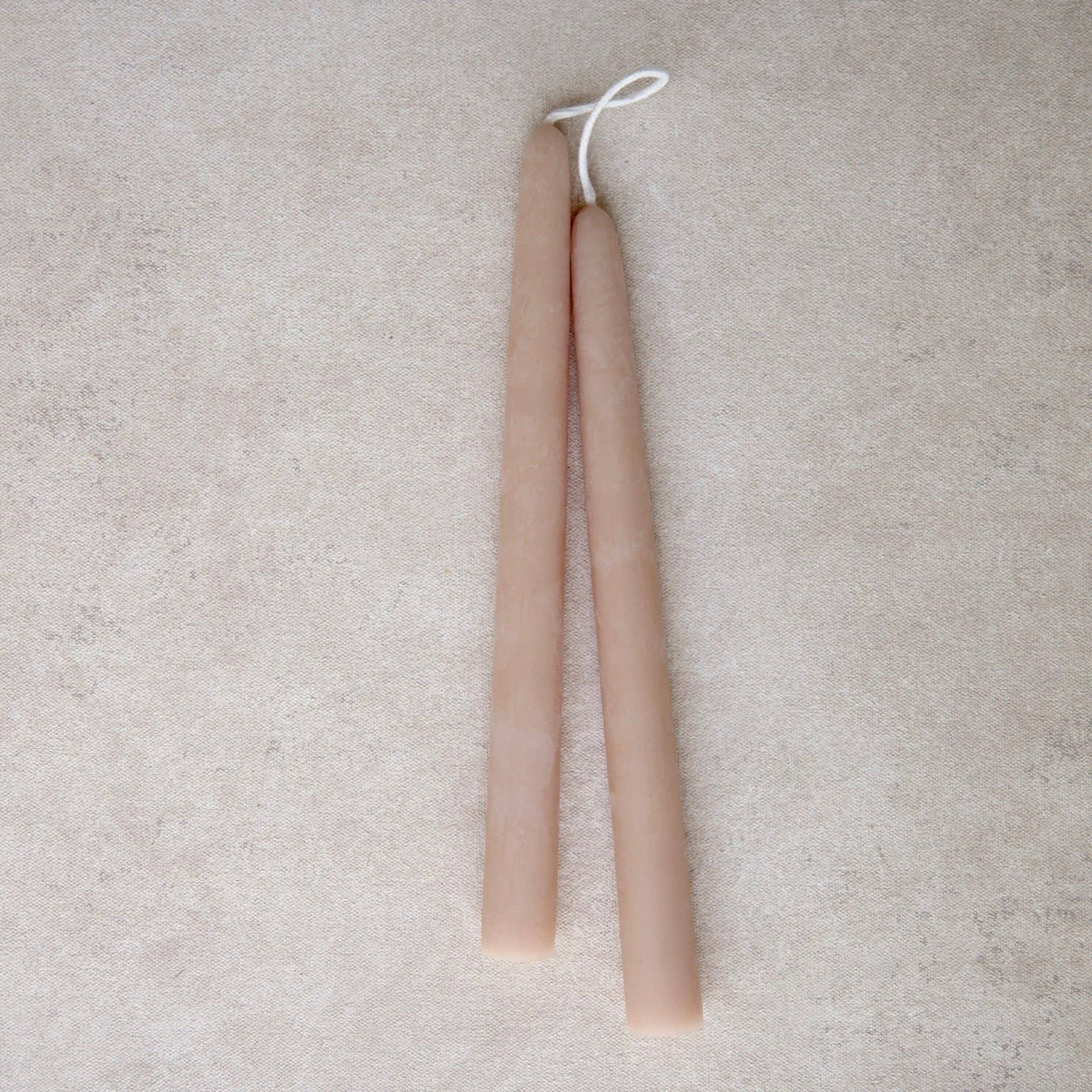 Two long taper candles in a light rose color, tied together with a white string at the top, displayed on a textured beige surface.