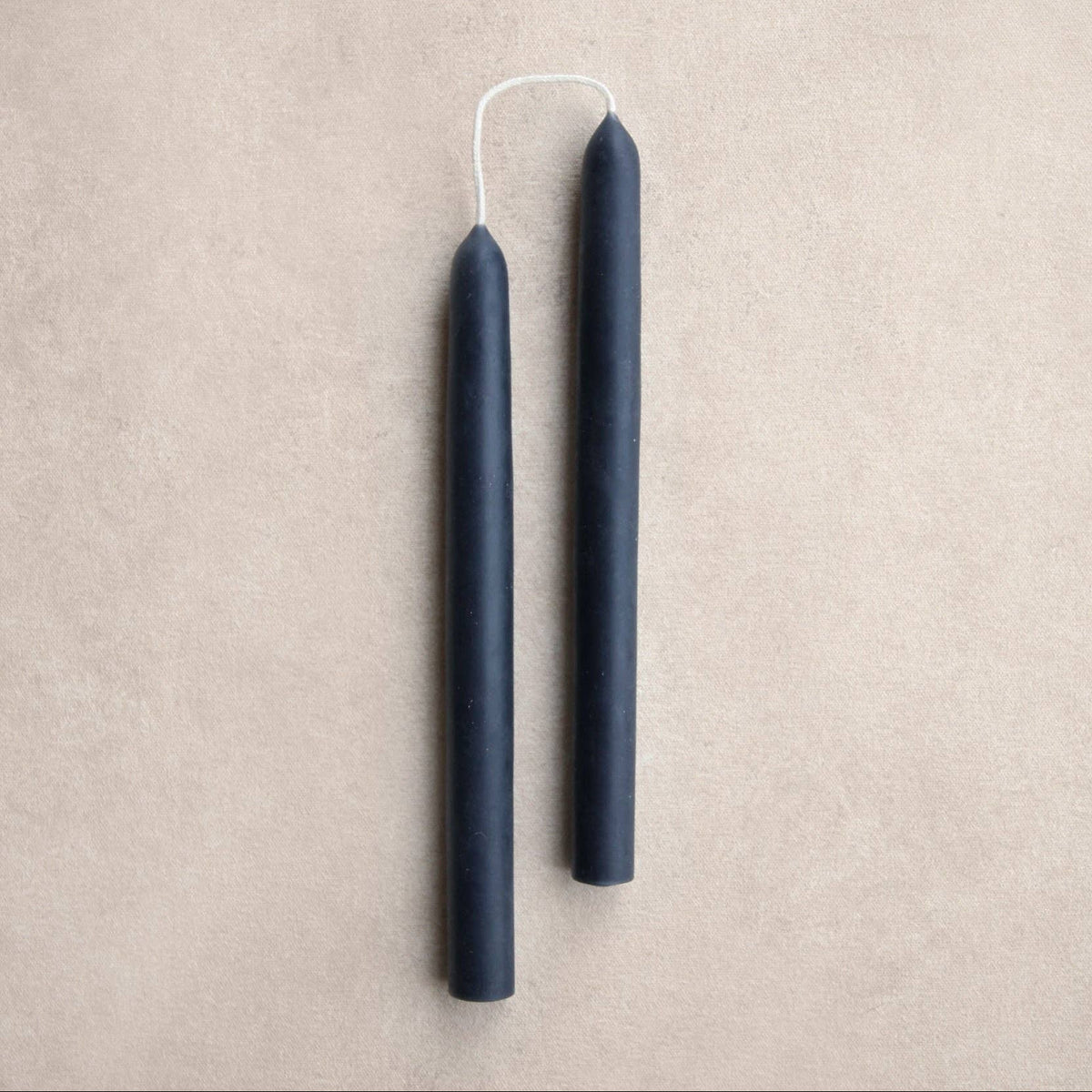Two ink color taper candles with white wicks, standing vertically on a textured beige background.