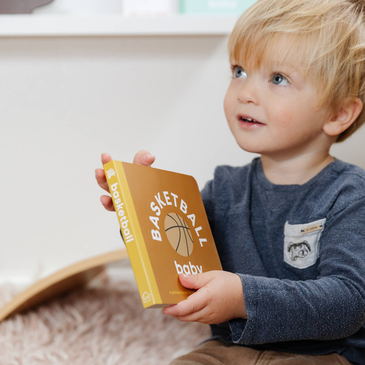 Child holding a book titled 'Basketball Baby' on a soft surface