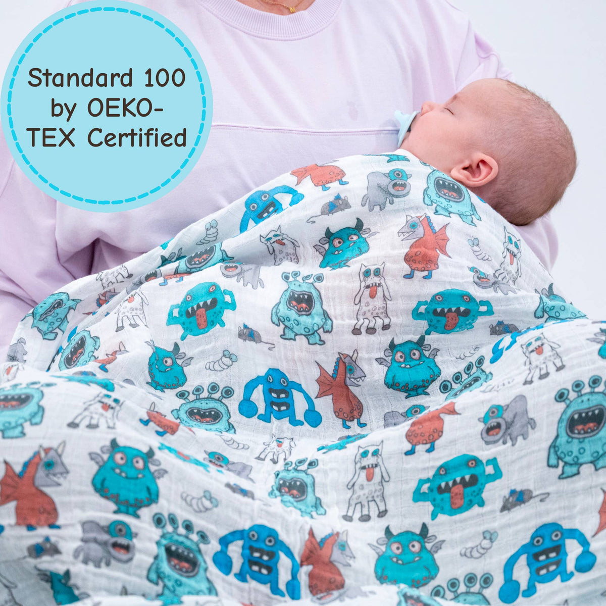 Baby wrapped in a blanket with cartoon monster pattern, held by an adult, with OEKO-TEX certification label.