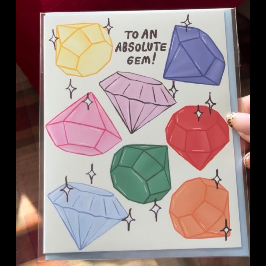 A colorful greeting card features the handwritten text "TO AN ABSOLUTE GEM!" in playful black lettering at the center, surrounded by nine sparkling, hand-drawn gemstones in various faceted shapes and vibrant colors: yellow, purple, pink, red, green, blue, and orange, each accented with little white starburst sparkles.