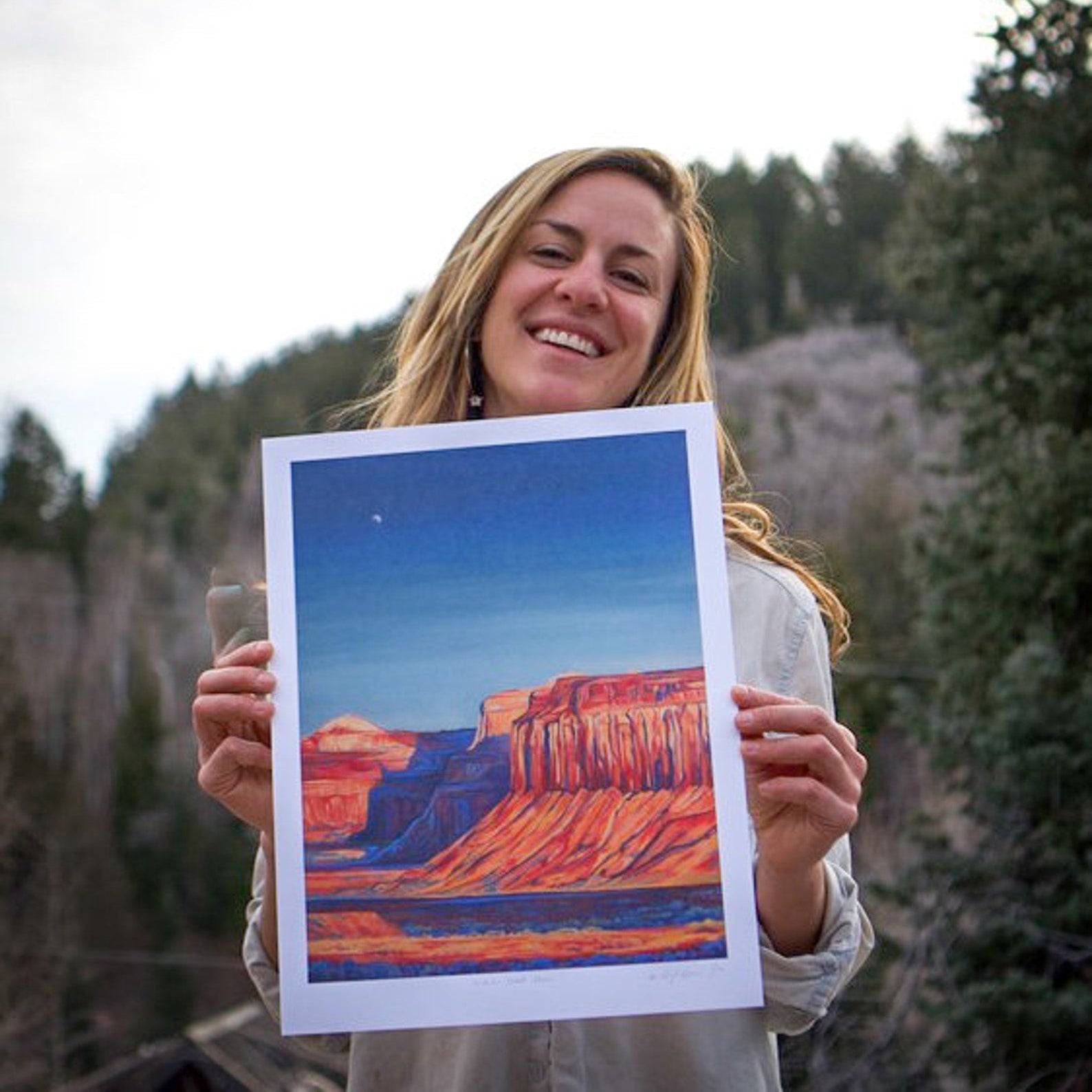 Woman holding a framed painting of a desert landscape in a natural setting.