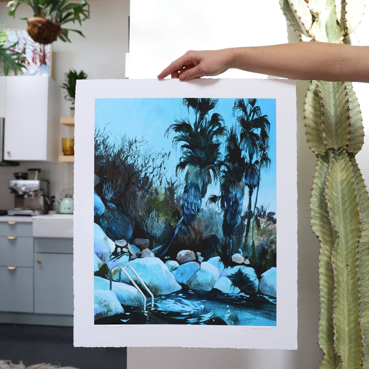 print held against a white wall with a cactus.