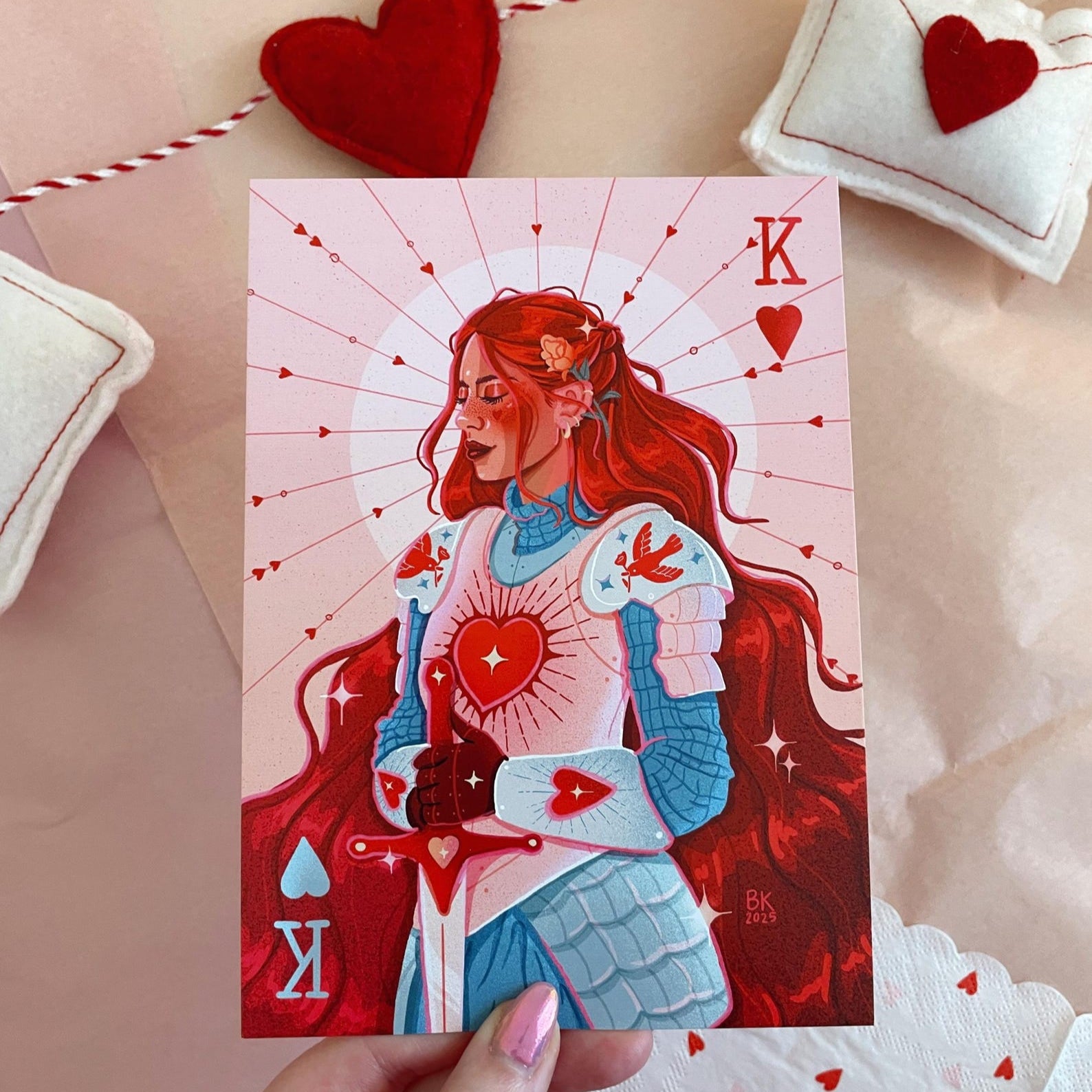 Knight of Hearts print pictures a fair knight dressed in light blue and pink armor with long flowing red hair. Print is held in front of pink tissue with a felt valentine garland in back ground.