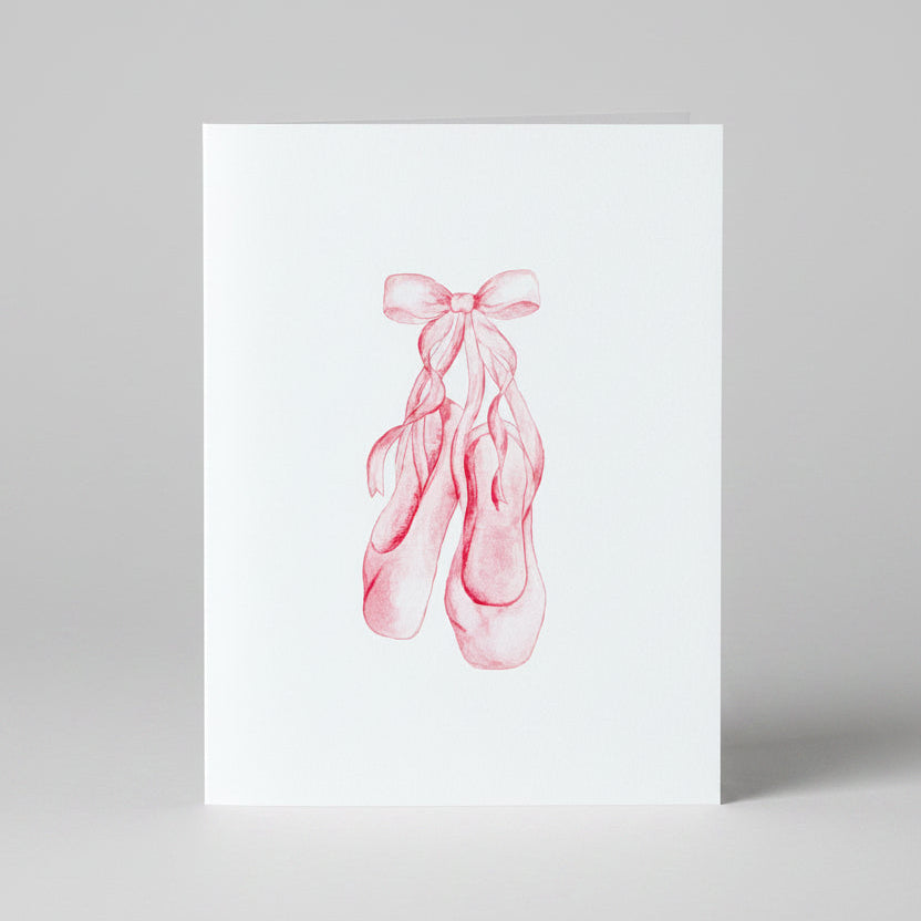 White card with pink ballet slippers tied into a bow. Shown against a wooden background.