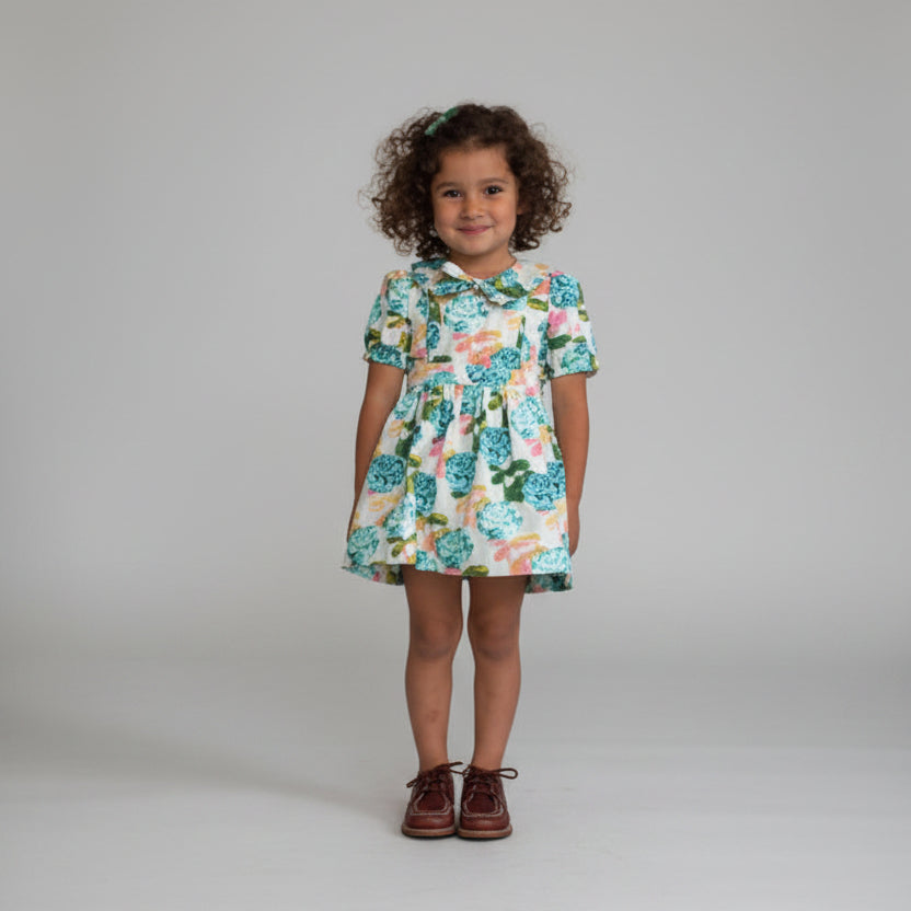Young girl wearing a floral dress standing against a white wall.