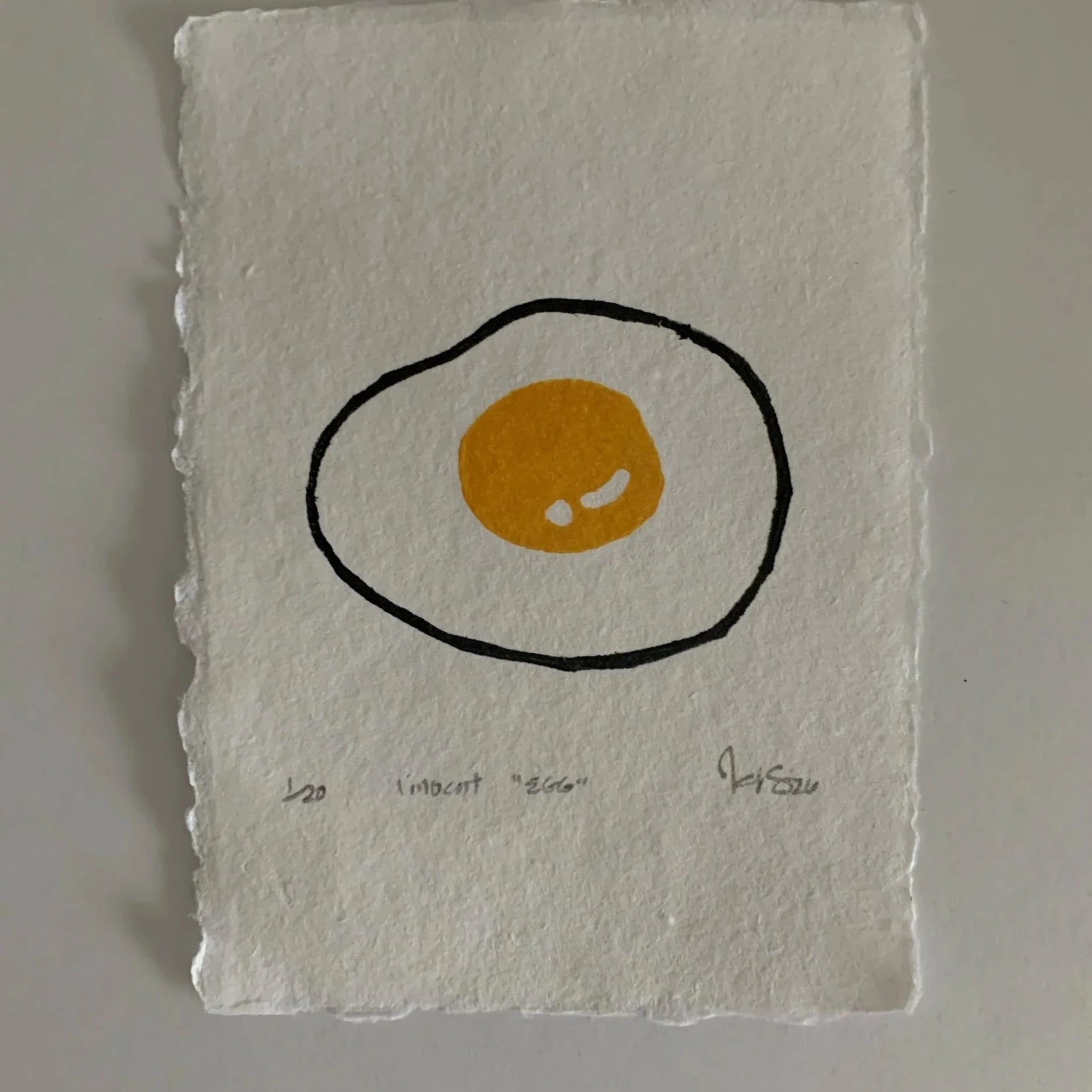 Linocut print on rough edge paper, depicting a sunny side up egg with a yellow yolk. Print signed by kelsi, with title and date. 