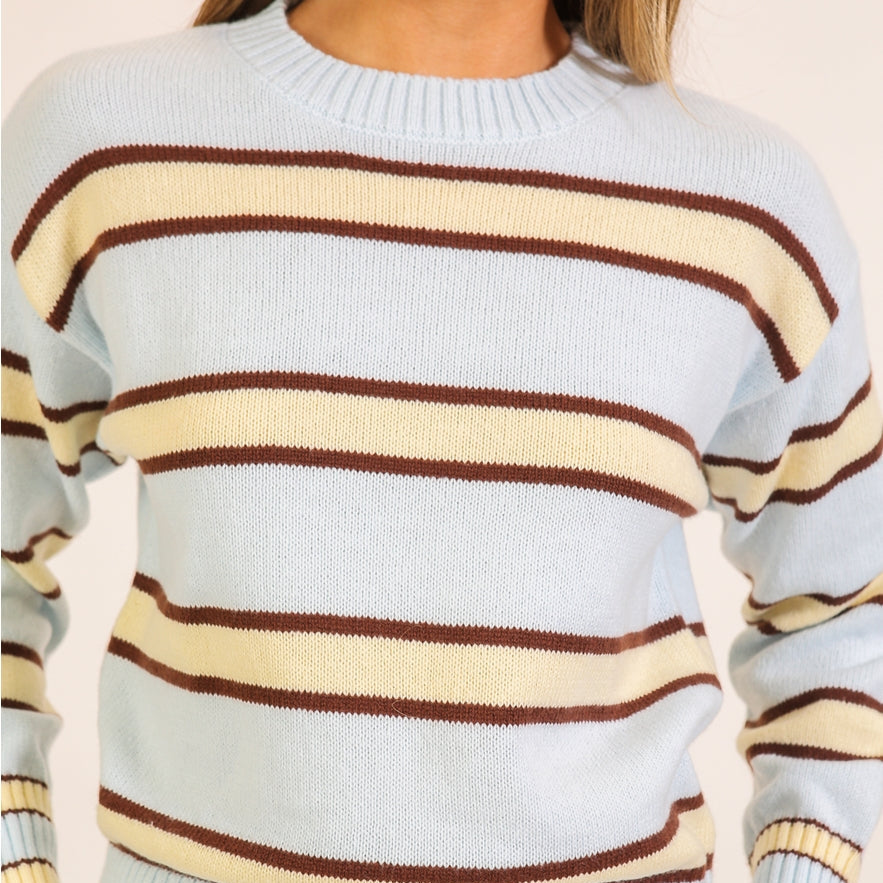 Woman wears a light blue crewneck sweater with horizontal yellow and brown stripes. 