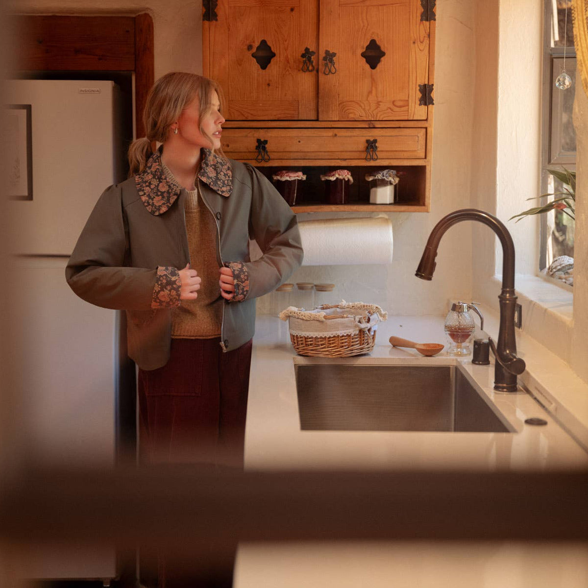 A woman stands in a warm, rustic kitchen wearing a green jacket with a patterned collar, a brown sweater, and a dark red pants. The scene is viewed through a dark wooden window frame, with wooden cabinets, a sink, and a window in the background.