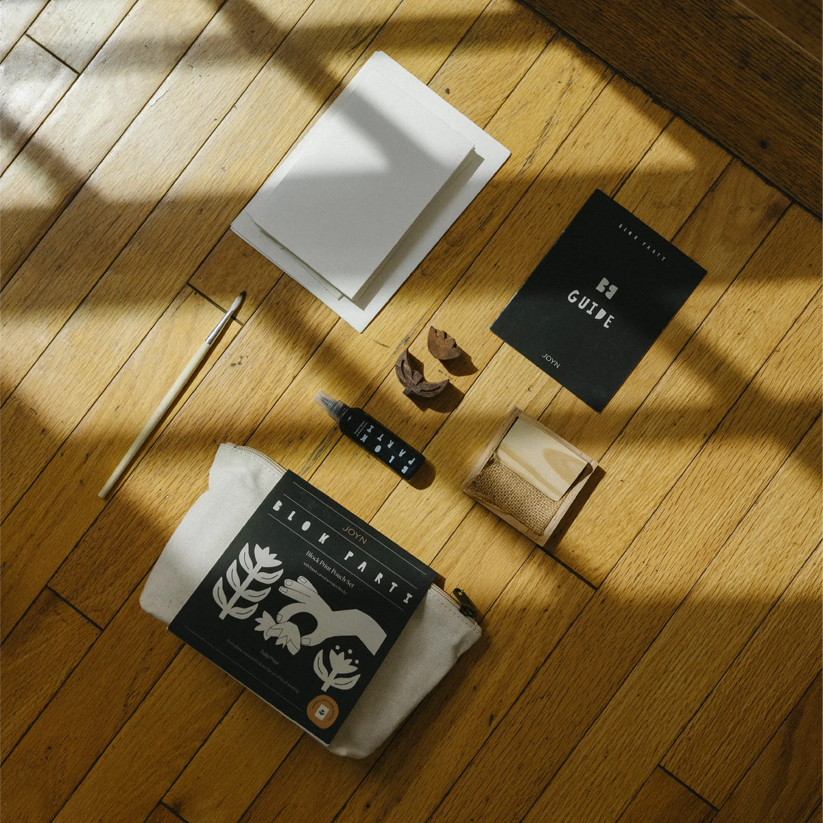 A styled flat-lay of minimalist stationery and accessories arranged on a warm wooden floor with strong sunlight shadows. Visible items include a black guidebook labeled 'GUIDE', a white envelope, a black pen, a small black pouch, a kraft paper bag, and a black-and-white book or notebook.