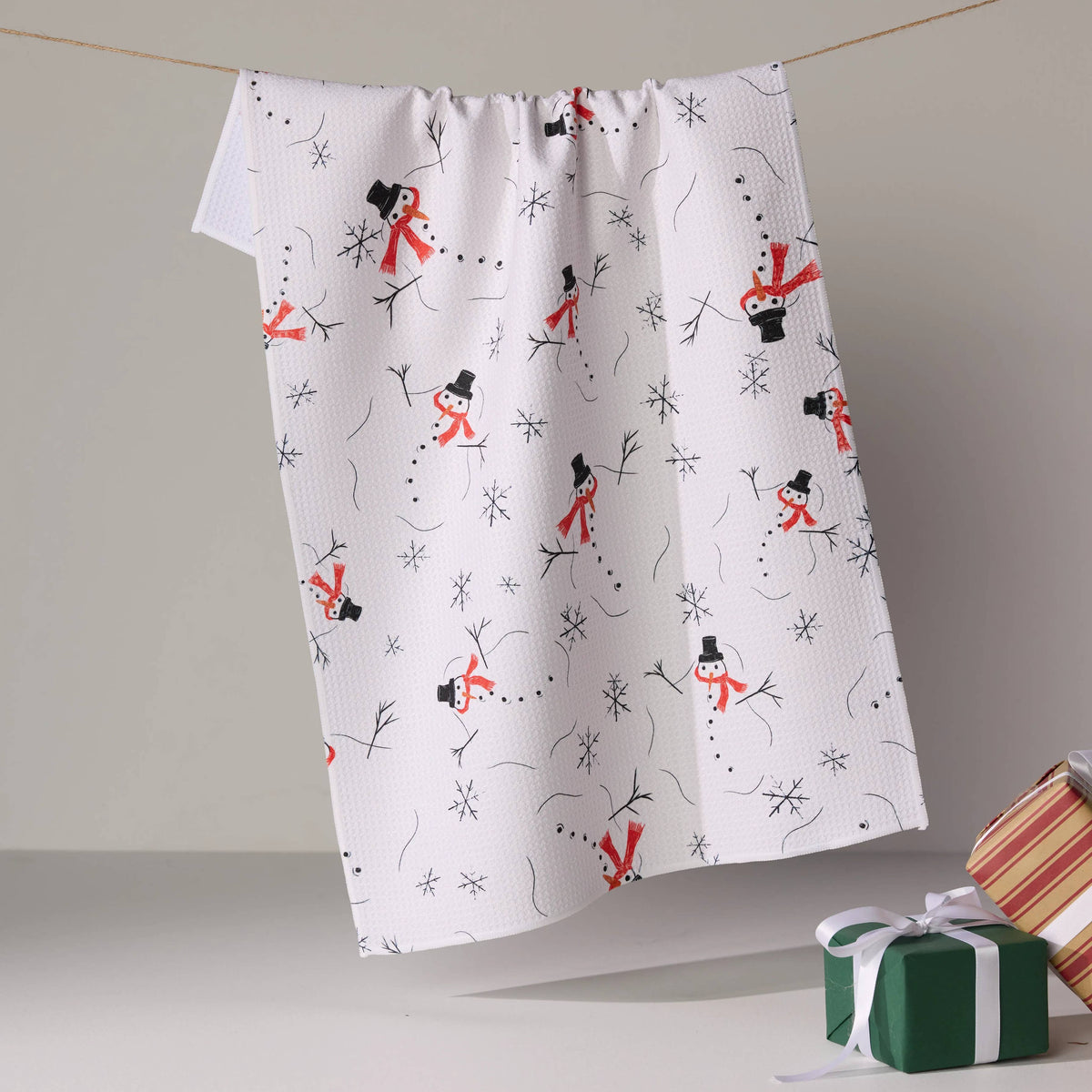 A white kitchen tea towel with a festive snowman print, hanging from a string in a studio setting with a green gift box in the foreground.