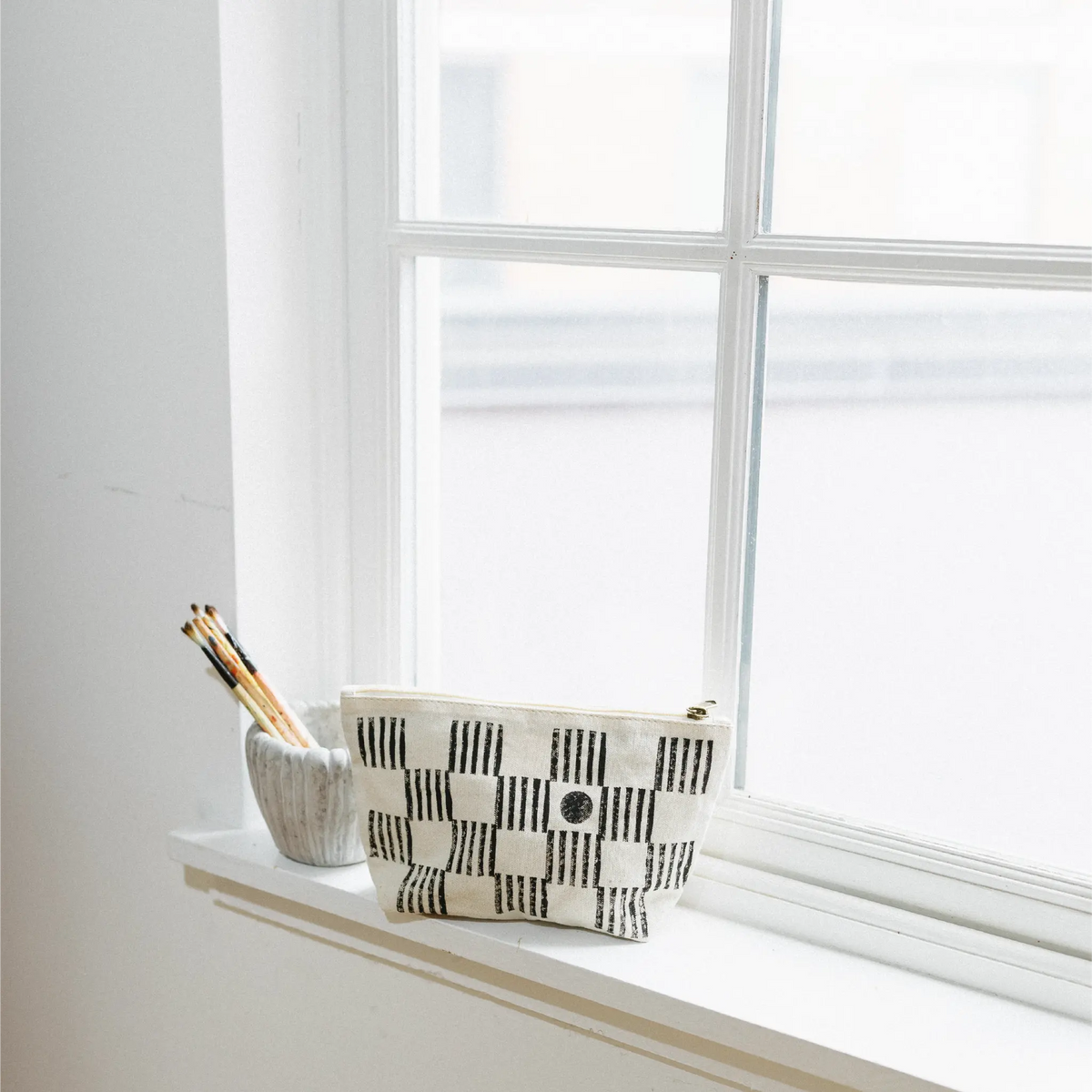 A small rectangular pouch with a black and white checkerboard pattern sits on a white windowsill next to a small gray cup holding pencils.