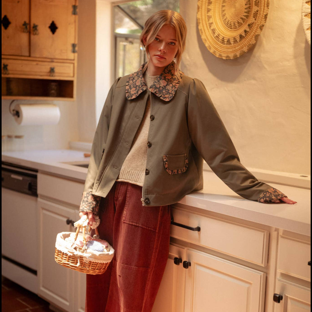 A woman stands in a warm, sunlit kitchen wearing a green button-up jacket with a floral collar, a cream knit sweater, rust-red wide-leg pants, and brown loafers. She holds a wicker basket against white cabinets and a tiled floor.