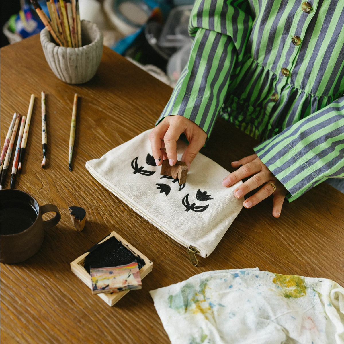 A person wearing a green and gray striped shirt is shown from the chest down, placing a small wooden stamp onto a cream canvas bag with black floral designs. The scene is set on a wooden desk surrounded by art supplies, including brushes, ink, and a cup of tea.