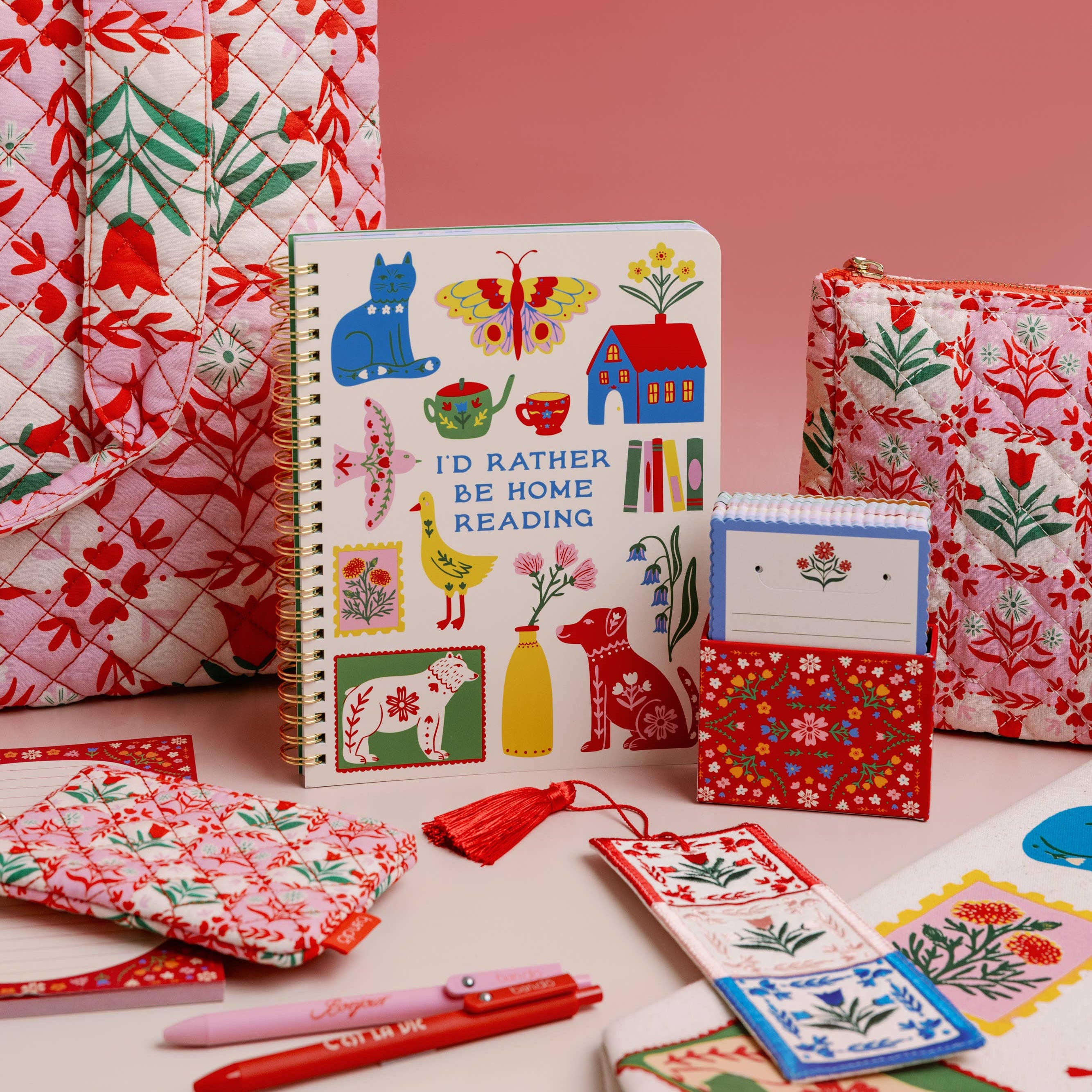 Collection of colorful stationery items including notebooks, cards, and a pen on a pink background.
