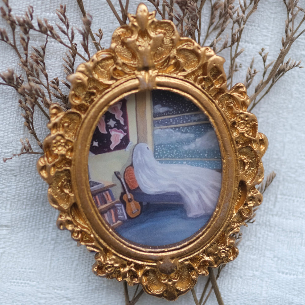A small, ornate gold frame holds a cozy illustration of a ghost on a bed, gazing out a window at a snowy night sky, with a guitar leaning nearby and books stacked on the floor. The frame is surrounded by delicate dried branches on a soft, light fabric background.