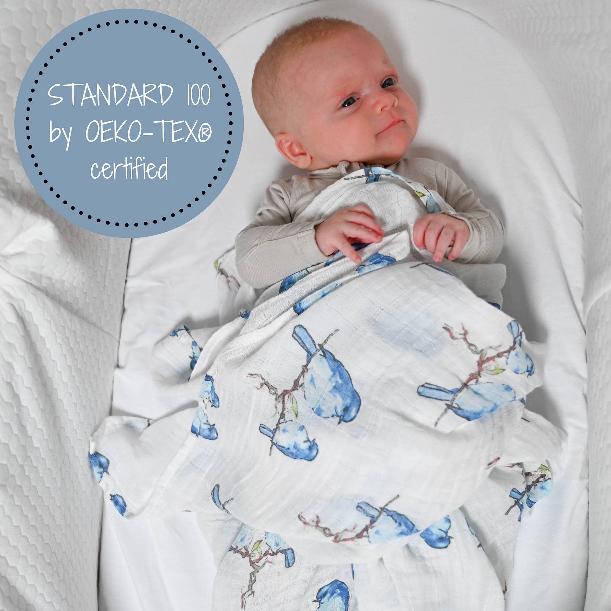 Baby wrapped in a blanket with blue bird pattern, lying on a white surface, with Oeko-Tex certification label.