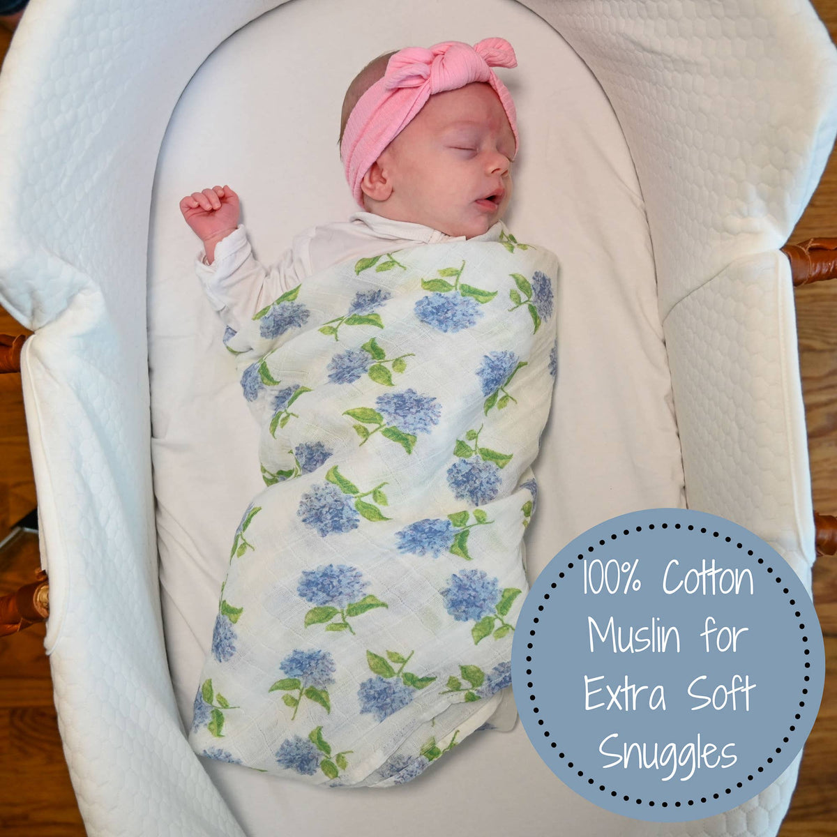 Newborn baby swaddled in a floral blanket with a pink headband, lying on a white cushion.