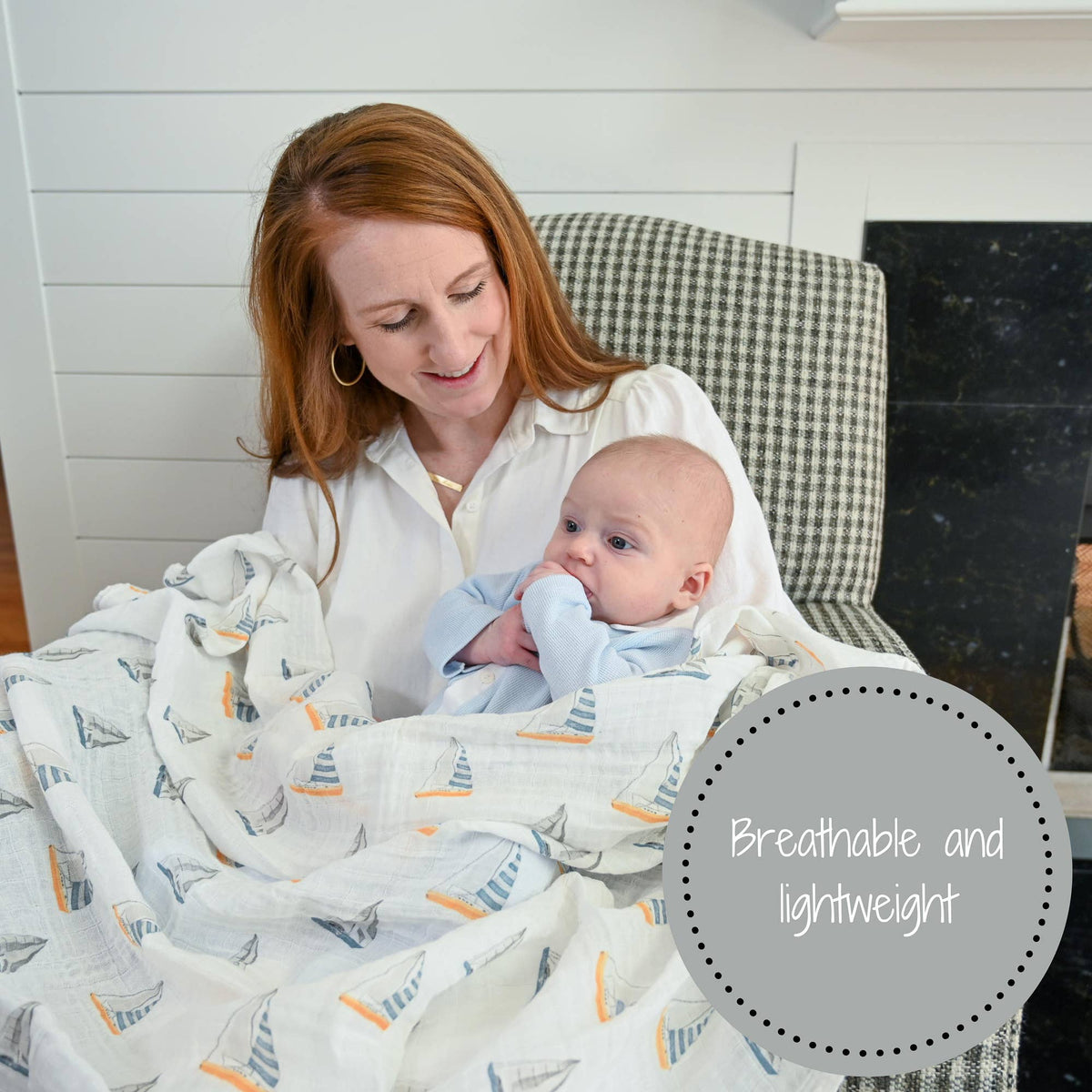 Woman holding a baby wrapped in a blanket with sailboat pattern, sitting on a checkered chair.