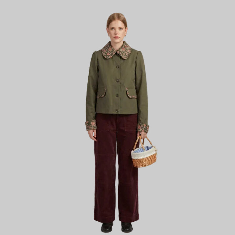 A woman stands outdoors in a wooded setting wearing an olive green jacket with a floral patterned collar and matching floral trim on the pocket, paired with dark red pants and holding a small woven basket.