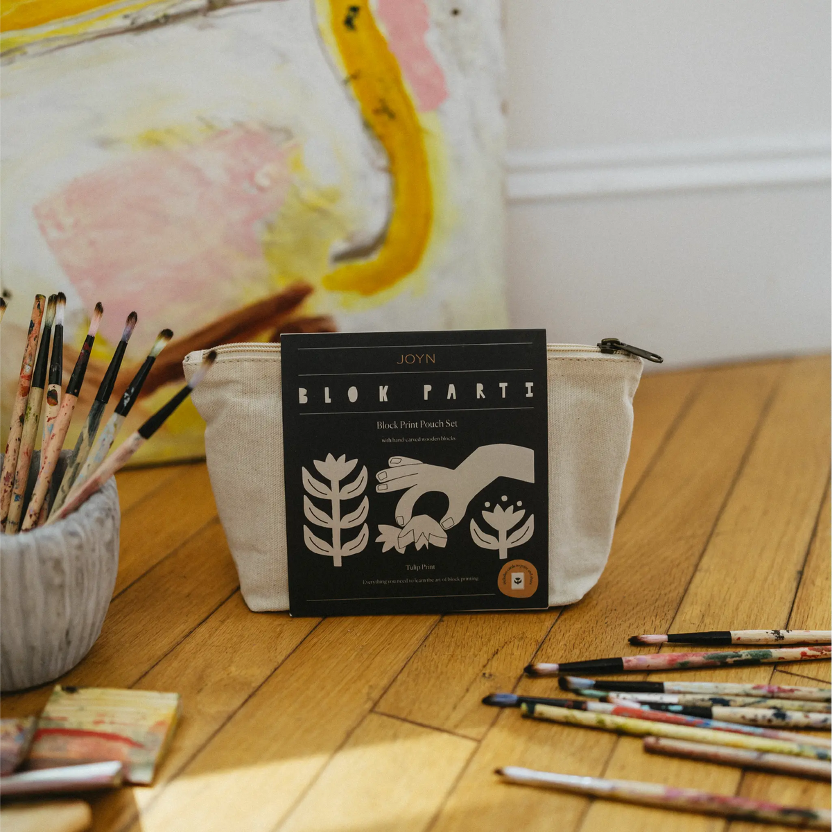 A canvas zip pouch with a black front label reading JOYN BLOK PARTY stands on a wooden floor, surrounded by paintbrushes and an abstract painting in the background.