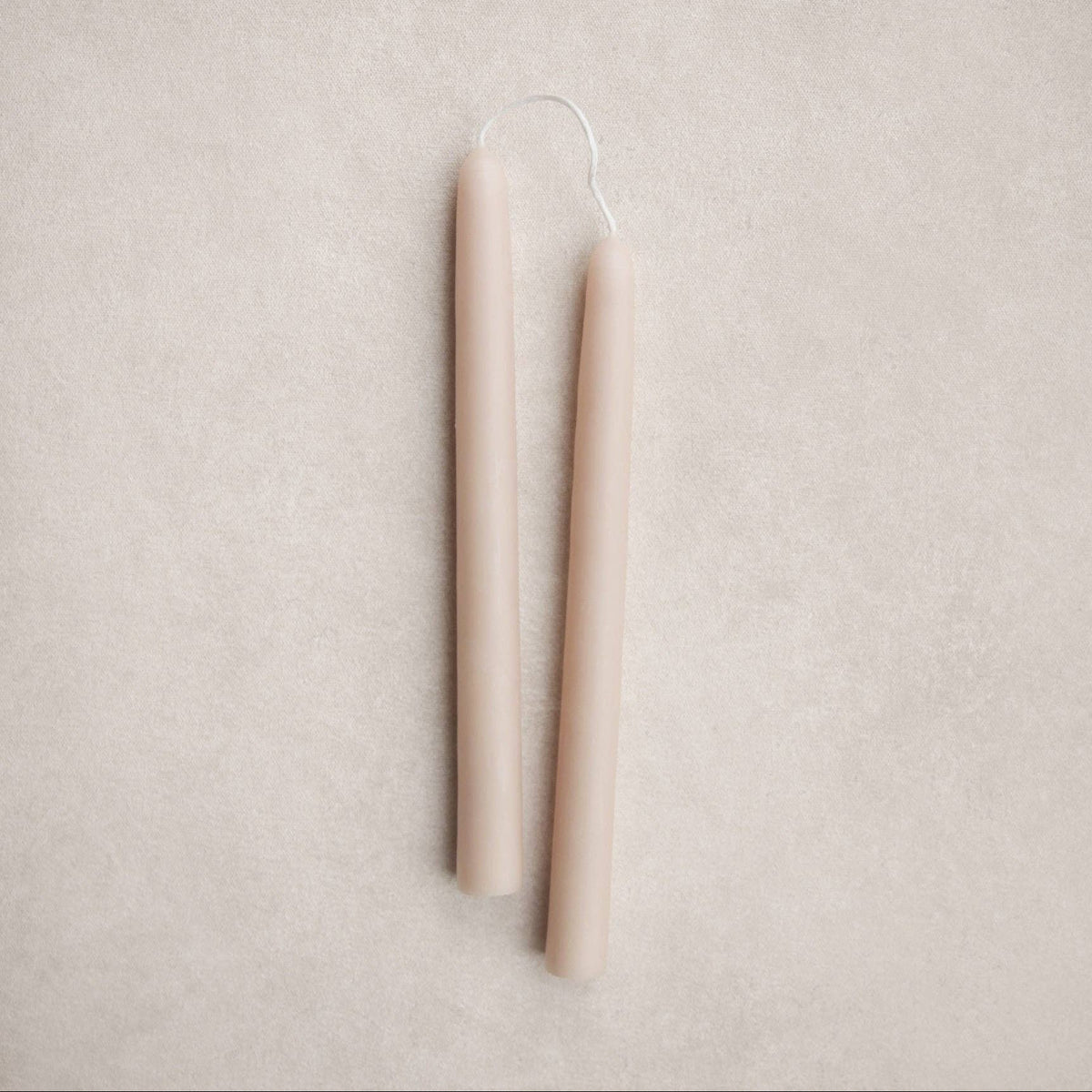 Two slim, tapered candles in a warm salt tone, shown upright on a textured light beige background.