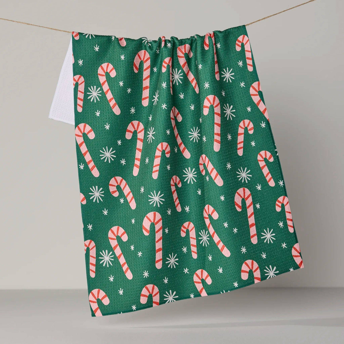 A green kitchen towel with a festive candy cane and snowflake pattern, hanging from a string against a neutral gray background.