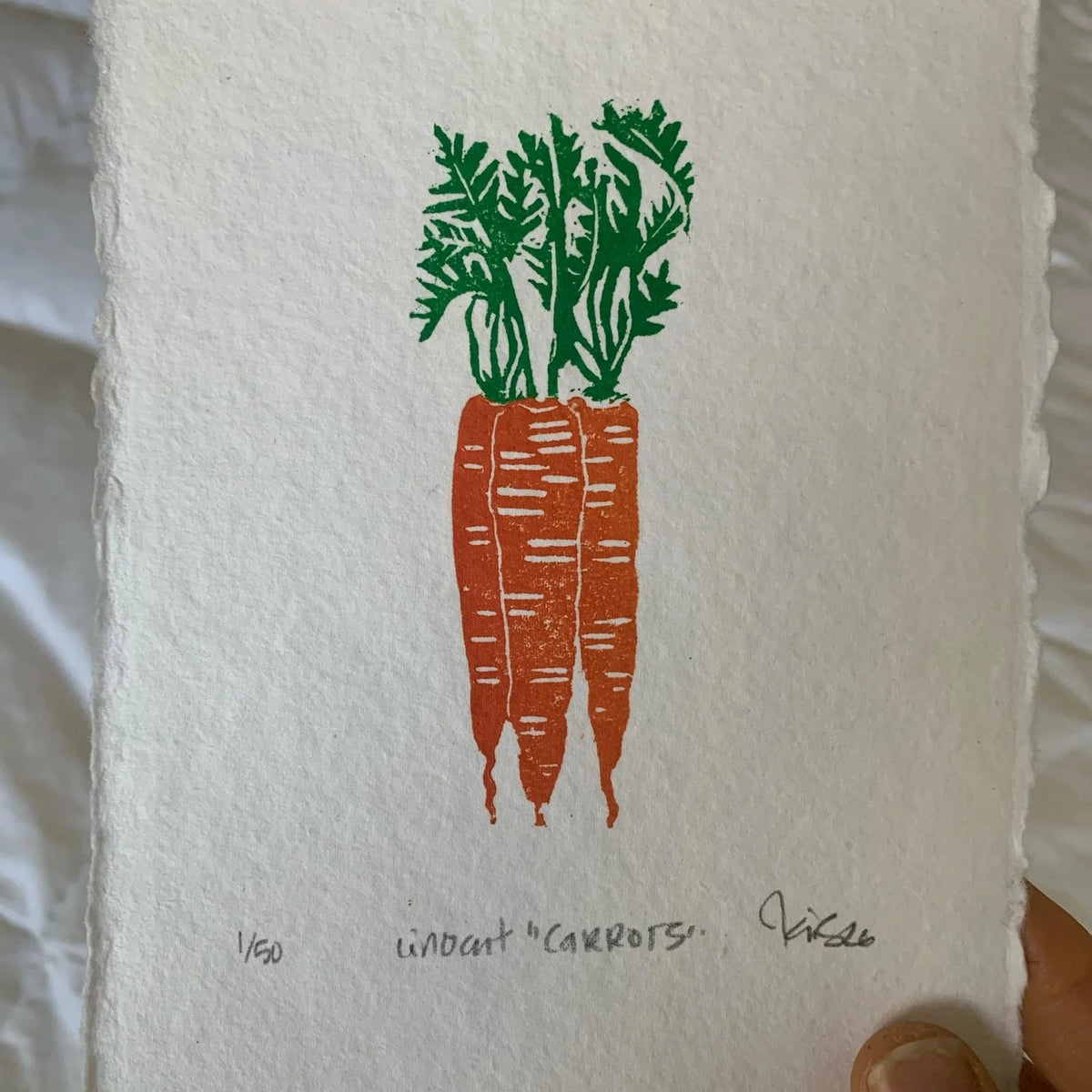 orange carrots with green tops in a bunch on 3. Linocut print done on rough paper, signed by Kelsi with title and date. Held in woman's hand against a white sheet.