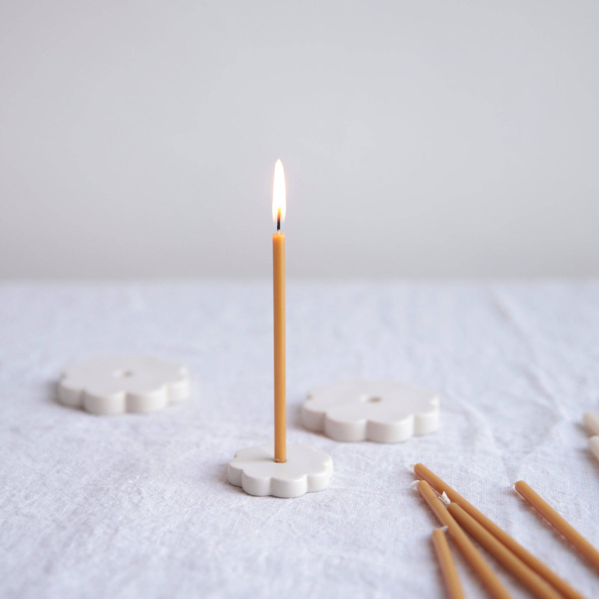 A single lit taper candle stands upright in a white flower-shaped holder on a textured white fabric surface, with additional unlit taper candles and more holders softly blurred in the background.
