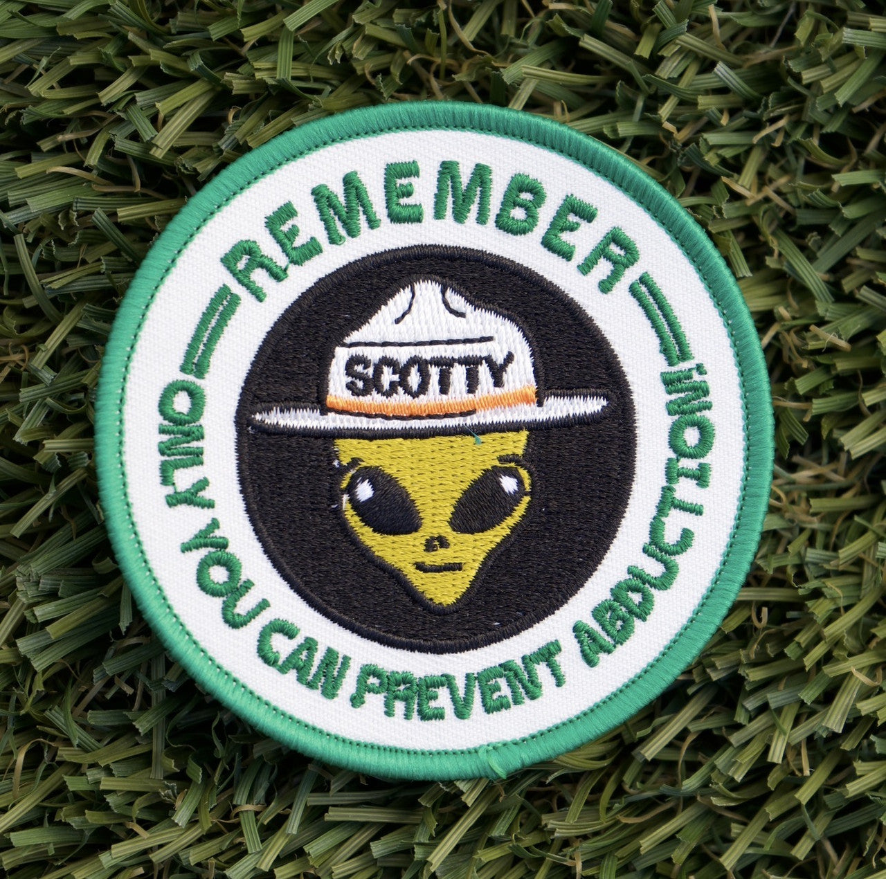 Round embroidered iron-on patch with an alien wearing a forest service hat with the words, "Remember, only you can prevent abduction" in green surrounding.