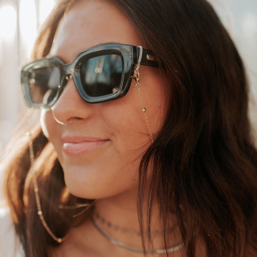 A close-up image of a person wearing sunglasses and a sunglasses chain with gold-colored accents.