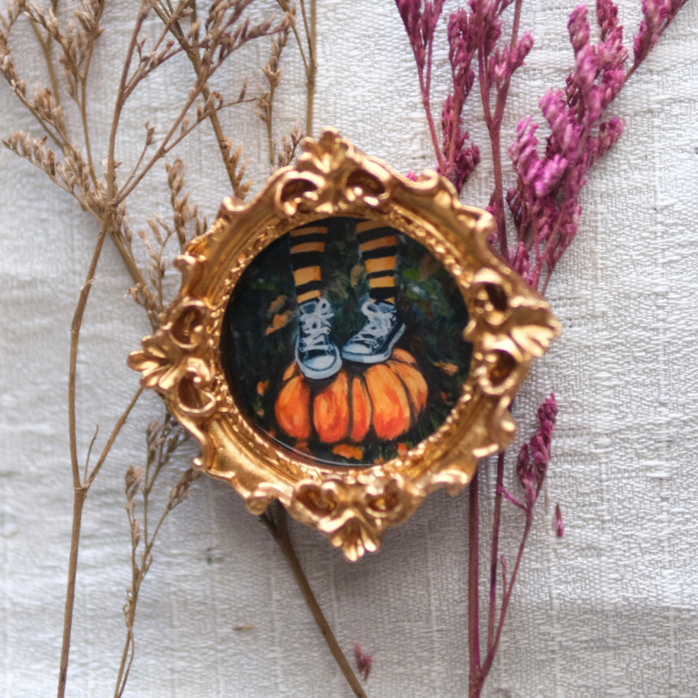 A small ornate gold frame holds an illustration of a pair of black-and-white striped legs wearing white high-top sneakers, standing atop two orange pumpkins in a dark, leafy setting. The frame rests on a soft white fabric background, encircled by delicate dried beige and purple branches.