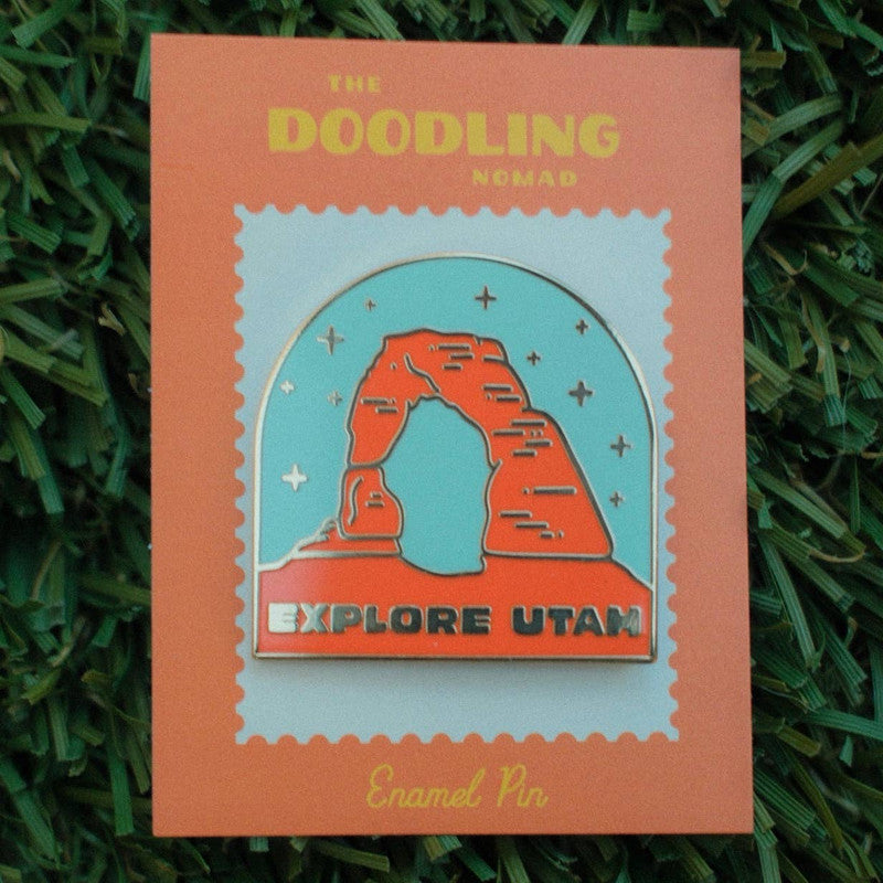 Enamel pin with a blue background and the Utah arches in orange color, with the words "Explore Utah" in gold.