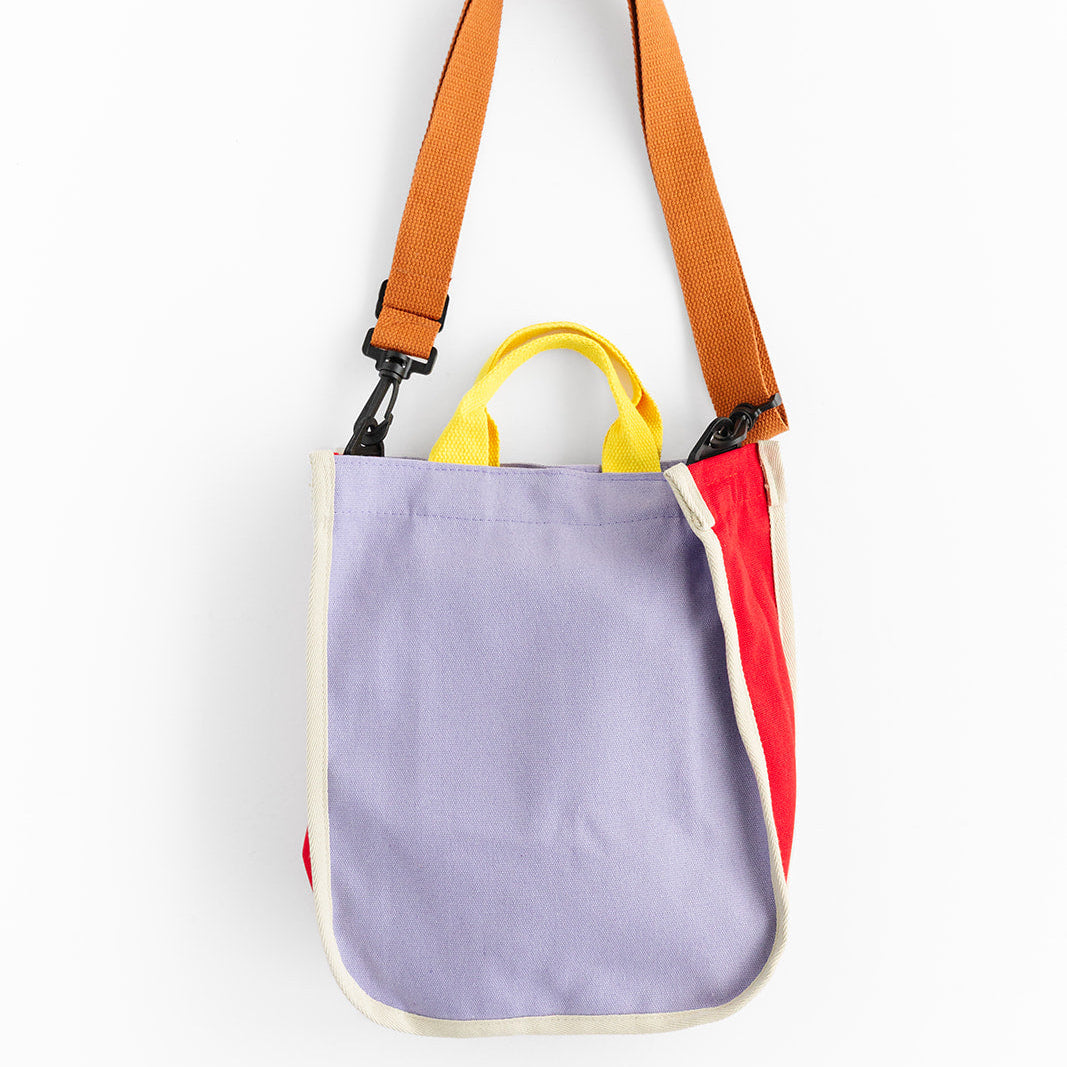 A colorful canvas tote bag features a lavender front panel, red side panels, cream trim, and yellow handles. An adjustable orange shoulder strap with black clips is attached to the bag against a white background. 