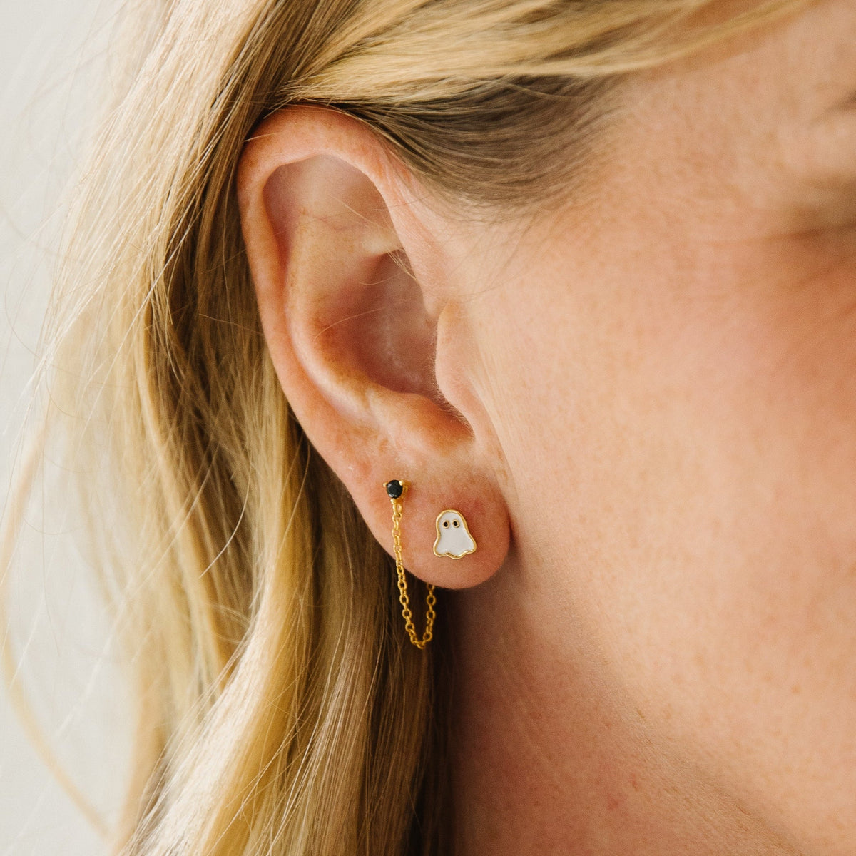 A close-up photo of a person wearing Halloween-themed ghost earrings, featuring a small white ghost stud with a gold outline paired with a delicate gold chain and black accent stud.