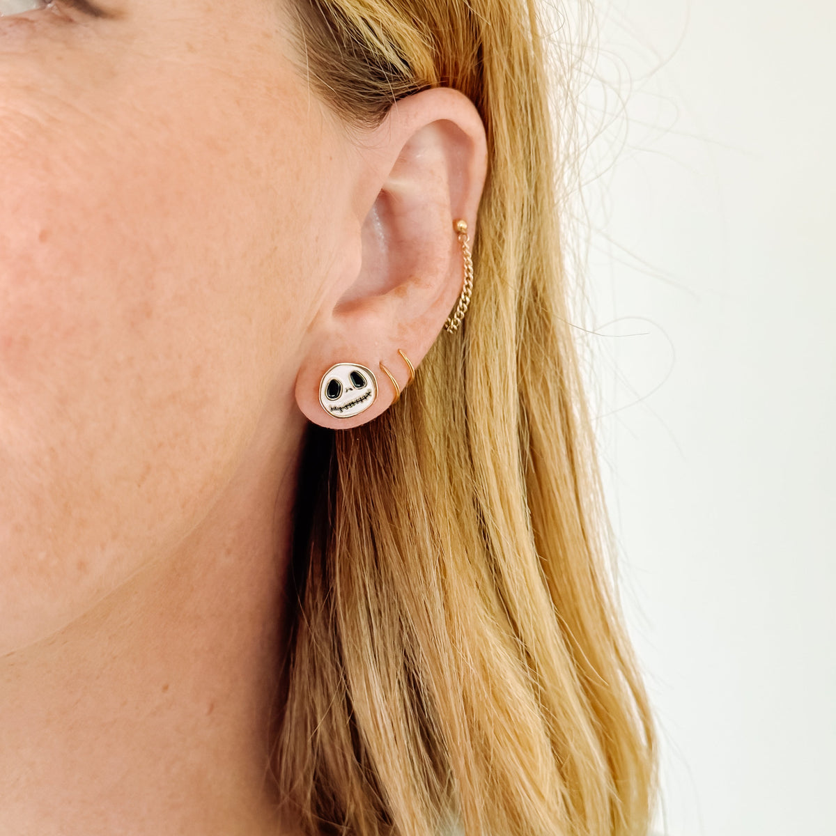 A close-up photo of a person wearing a gold-trimmed skull character stud earring with large black eyes and a stitched smile, paired with a delicate gold ear chain.