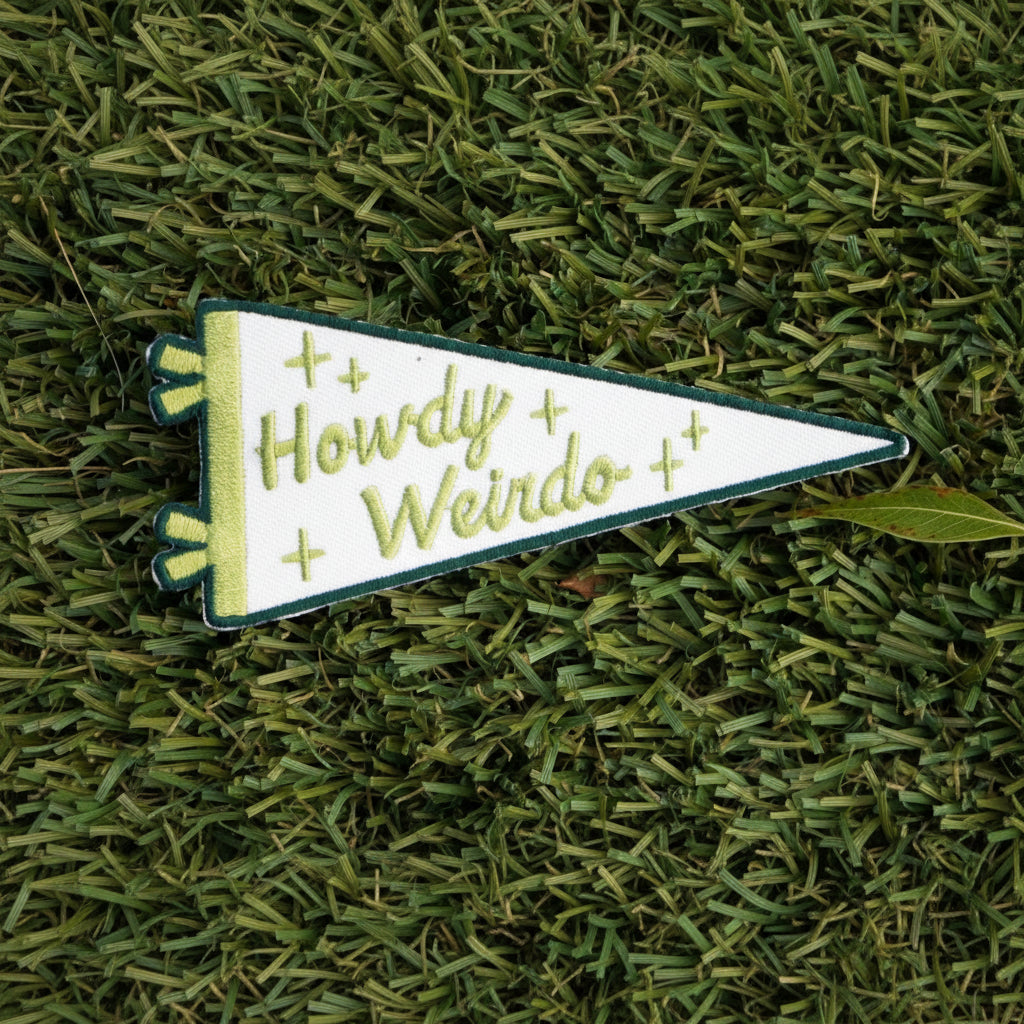 Iron-on  embroidered patch triangle shaped, with a white background and green words that say "Howdy Weirdo".