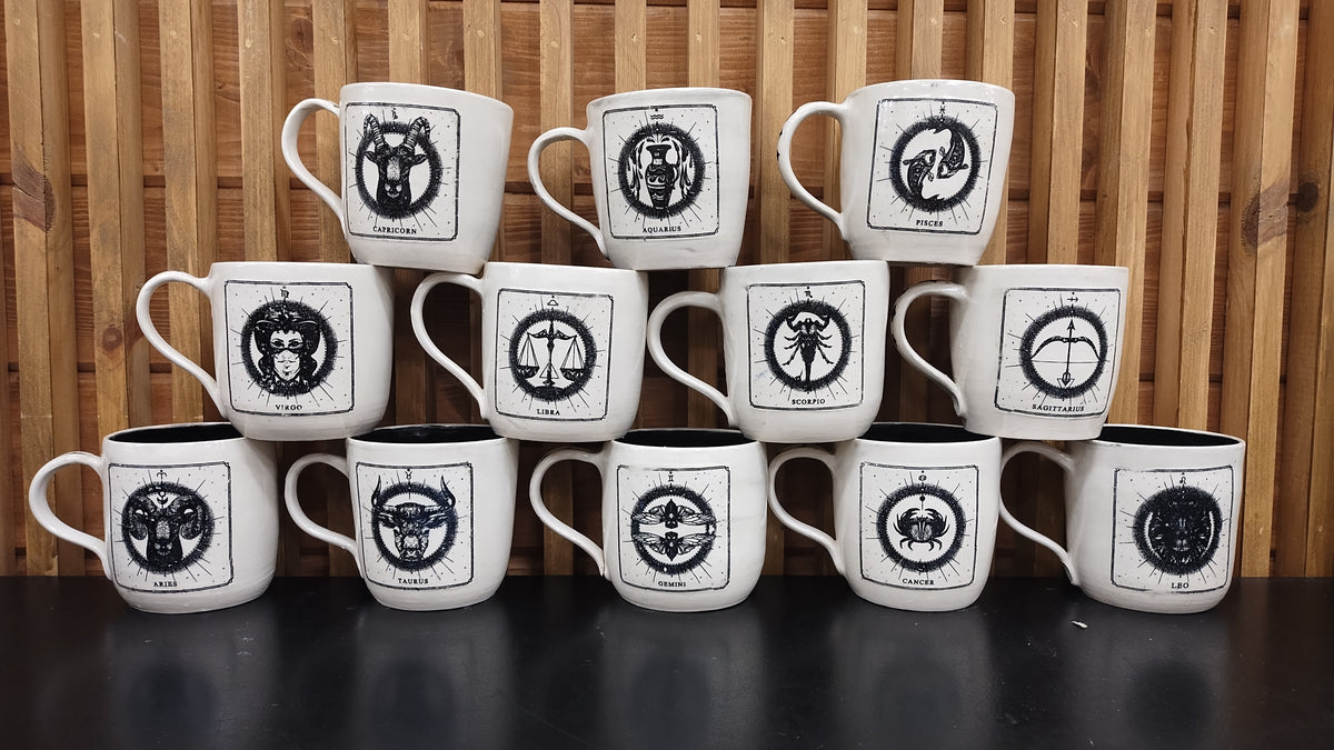 Warm beige stoneware mugs with a black zodiac sign design for each mug. 