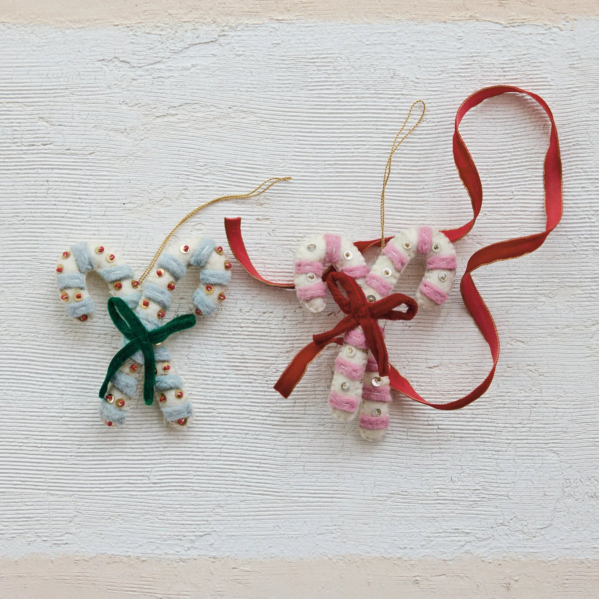 Two handmade  felt holiday ornaments shaped like candy canes, made from white and pink striped fabric with small embroidered details, hanging from thin twine with red ribbons, displayed on a textured off-white background.