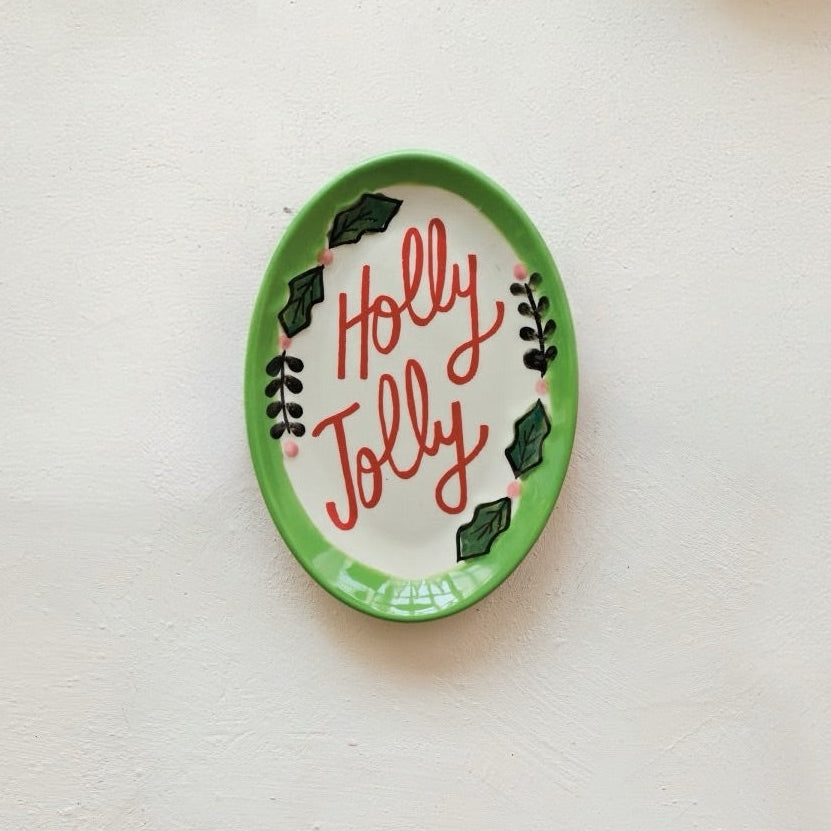 An oval ceramic dish with a green rim and the words 'Holly Jolly' written in red script, decorated with green holly leaves and small pink berries, displayed on a textured off-white wall.