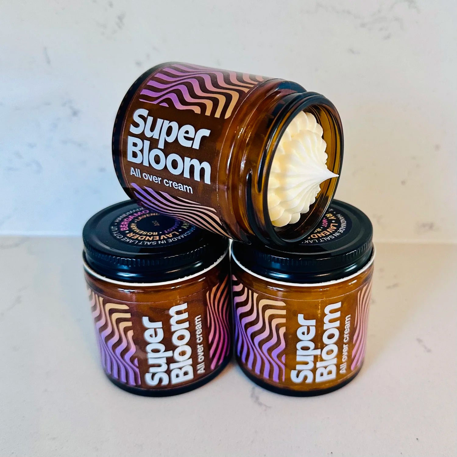 A stack of four small amber glass jars of "Super Bloom" all-over cream sits on a marble surface, with one jar tipped forward to show its fluffy white whipped texture inside and a black lid with colorful wavy purple-pink-white patterns. The jars feature matching retro-inspired labels in swirling pastel gradients, gold or black metal lids (one with a "Bergamont • Lavender • Rosemary" sticker), and are casually arranged to highlight the product's luxurious handmade appearance.