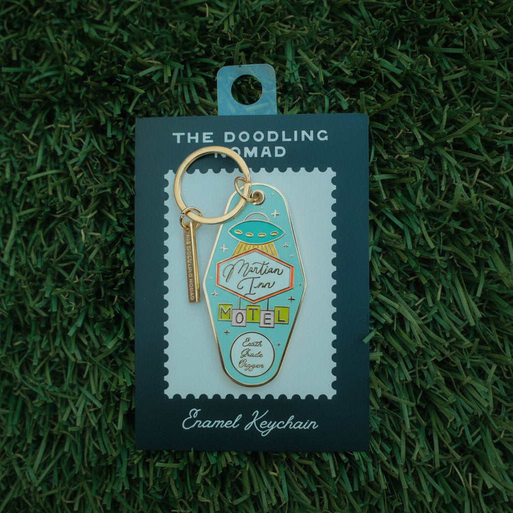 Diamond-shaped enamel keychain in turquoise color, with an alien ship and a sign that says "Martian Inn Motel" and "Earth, Grade, Oxygen".