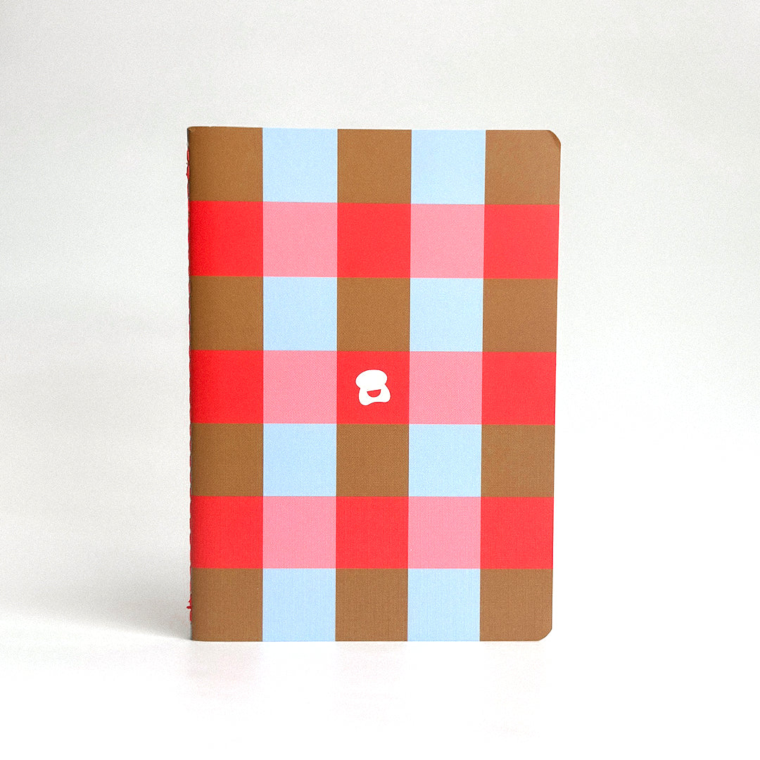A notebook with rounded corners features a bold plaid pattern in brown, light blue, pink, and red. A small white smiley logo is centered on the cover. 