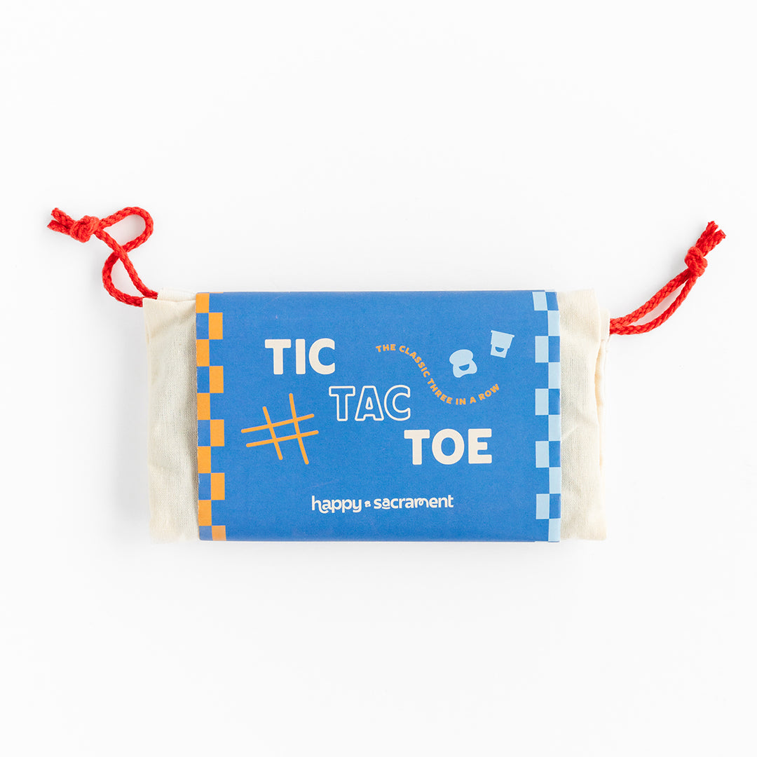 A blue and orange Tic Tac Toe game set is displayed in a small cream-colored drawstring pouch with red cords. The colorful package features the words “TIC TAC TOE” and “happy + sacrament” on a white background.
