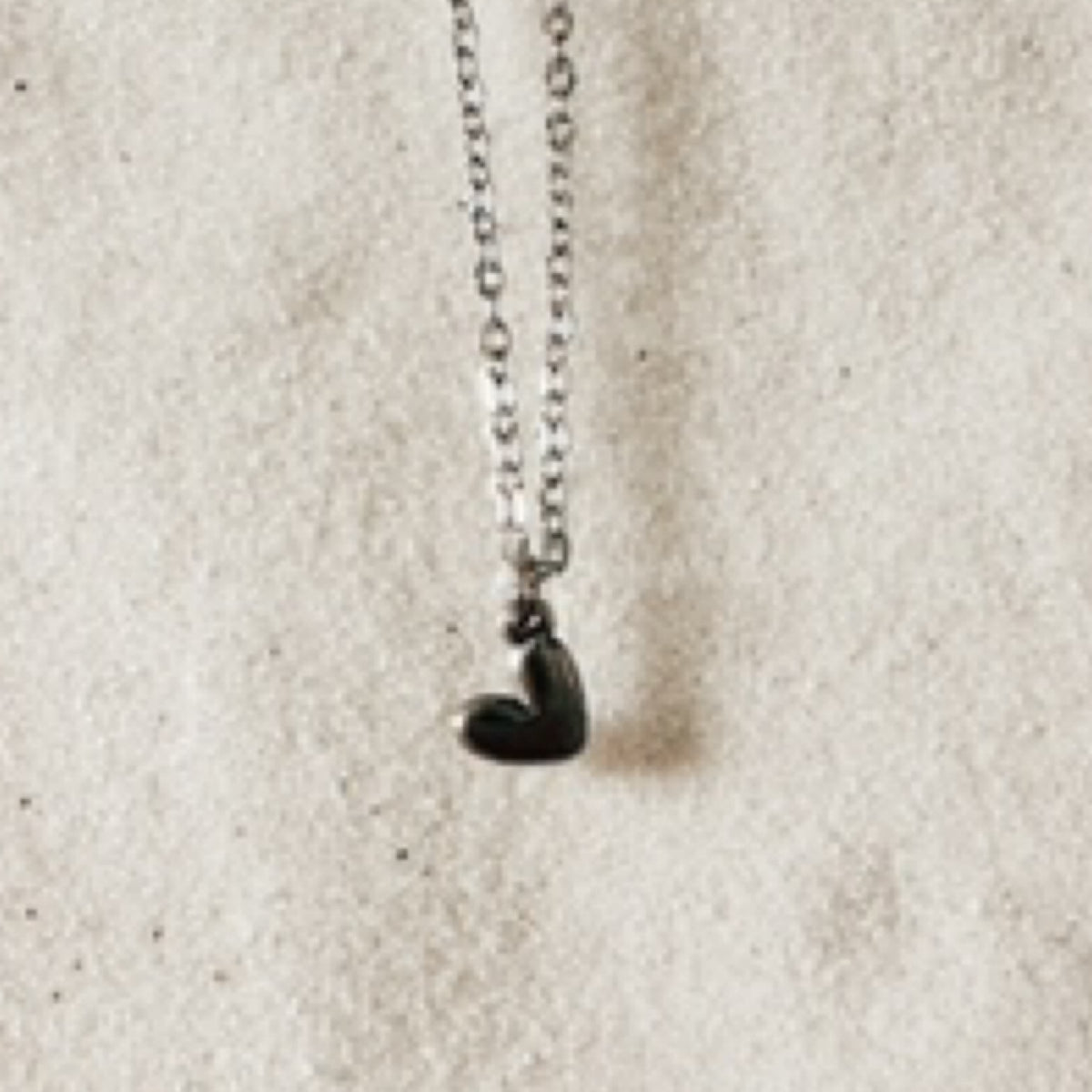 Delicate silver chain with a small flat heart pendant hanging on a tilt. Pictured against white sand.