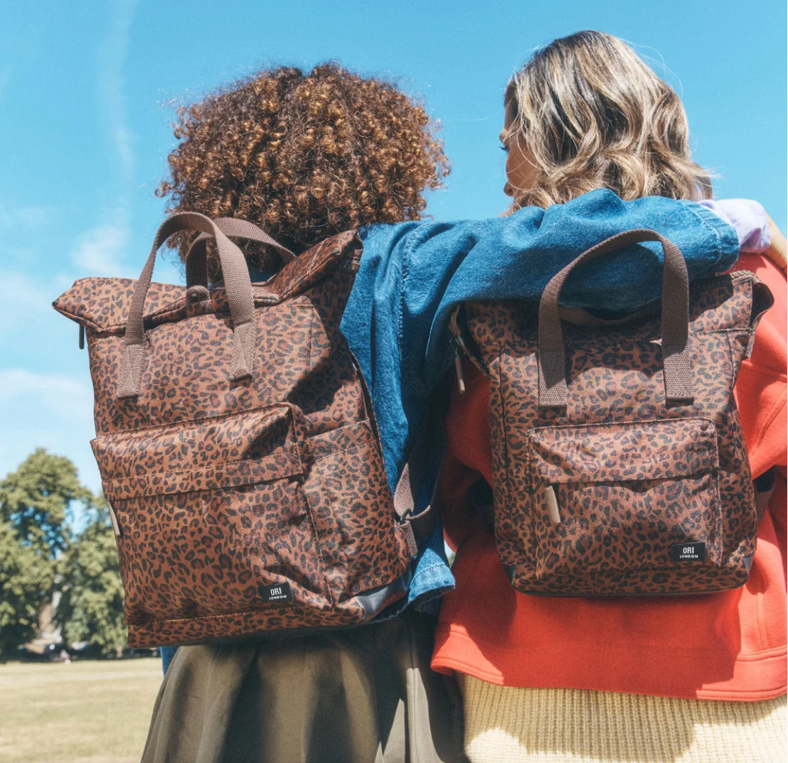 Two people seen from behind wearing matching leopard-print backpacks outdoors under a blue sky. The backpacks have a brown and tan animal print with dark brown handles and trim, and small black brand patches near the bottom corners.