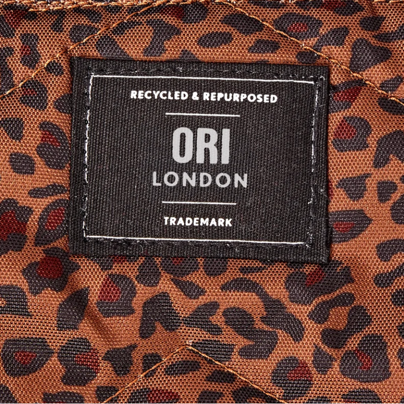 Close-up of a brown and black leopard print fabric with a central black brand patch reading ORI LONDON, plus two dark snap buttons at the top.