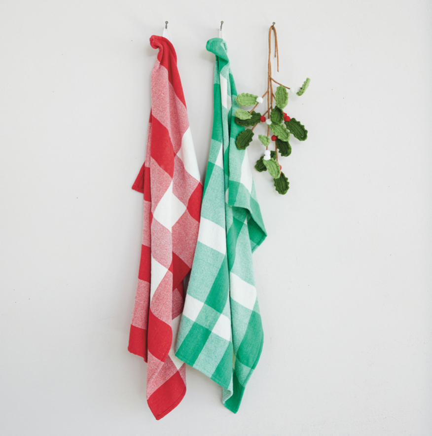 Two checkered kitchen towels hanging from metal hooks on a white wall, one red and white and one green and white, with a small green leafy sprig hanging on the right.